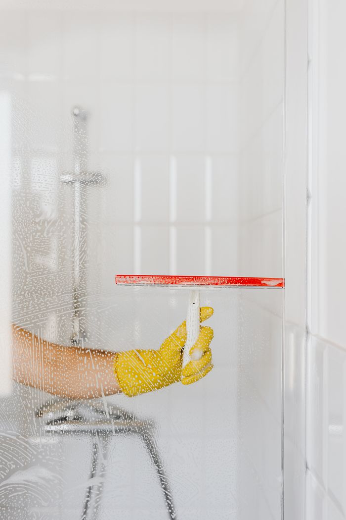 about-01 Crop anonymous person in yellow rubber glove cleaning glass shower unit with glass wiper and detergent against white tile wall