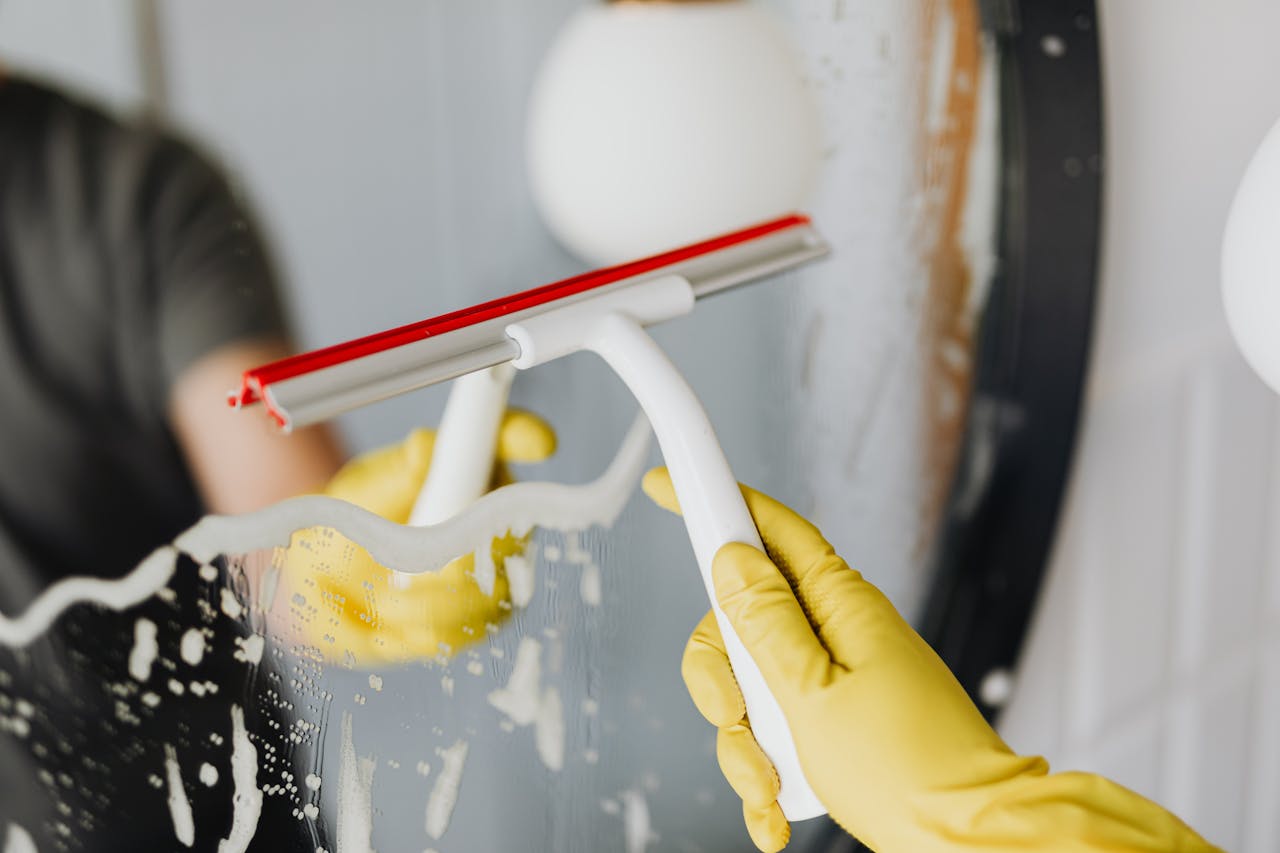 about-03 Faceless person in yellow rubber gloves using scraper to remove soapy water from mirror in bathroom