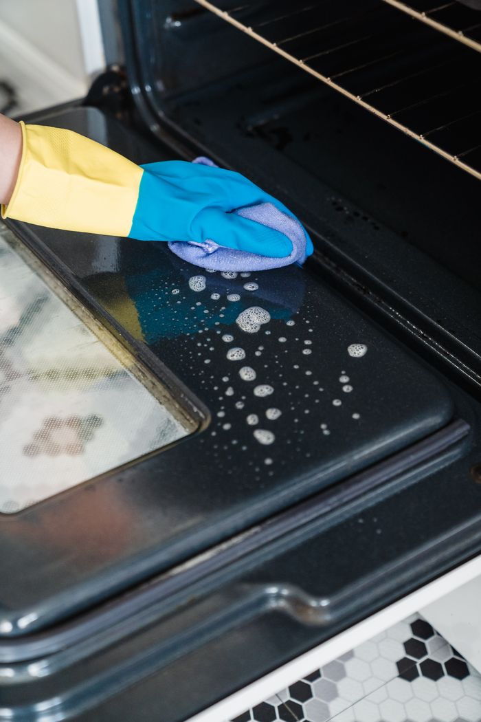 our-story-01 Close-up of cleaning an oven door with gloves and detergent in a kitchen.